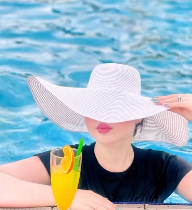A woman in a large white sun hat and black top sits by a pool holding a glass of orange juice garnished with an orange slice.