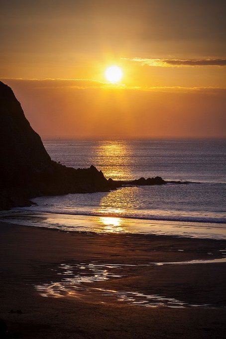 A scenic photograph of a sunset over the ocean, with golden light reflecting on the water and wet sand at a beach bordered by dark cliffs.