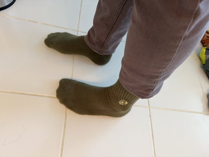 The image shows a person wearing green socks, standing on white tiles with their feet positioned at the bottom of the frame. The photo appears to be taken from an angle that captures the person's legs and shoes, providing a clear view of their footwear.