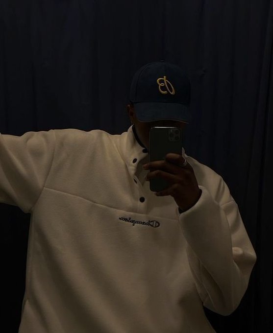 A person takes a mirror selfie wearing a navy Champion cap and a cream-colored Champion fleece pullover, obscuring their face with a smartphone.