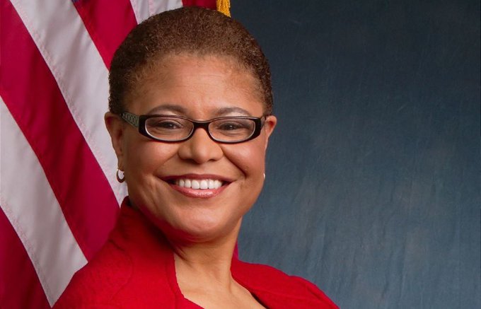 An official portrait of Congresswoman Karen Bass, a woman with short hair and glasses wearing a red blazer, smiling in front of an American flag.