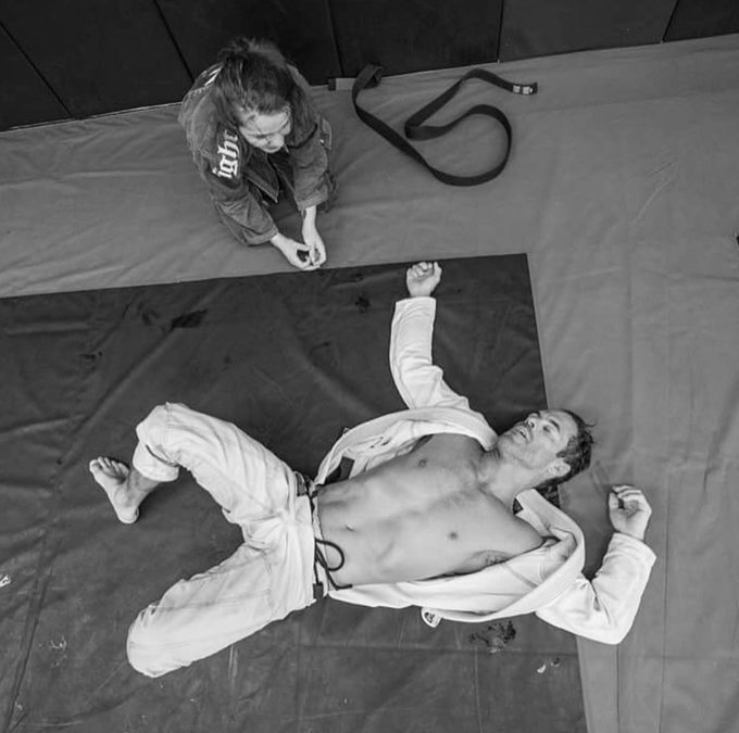 A black-and-white overhead photo shows a man in a white gi lying defeated on the mat while a young girl in a black gi sits nearby tying her belt, with the word "light" visible on her uniform—suggesting the "trading" in 2026 may not refer to markets but rather a humbling martial arts lesson.