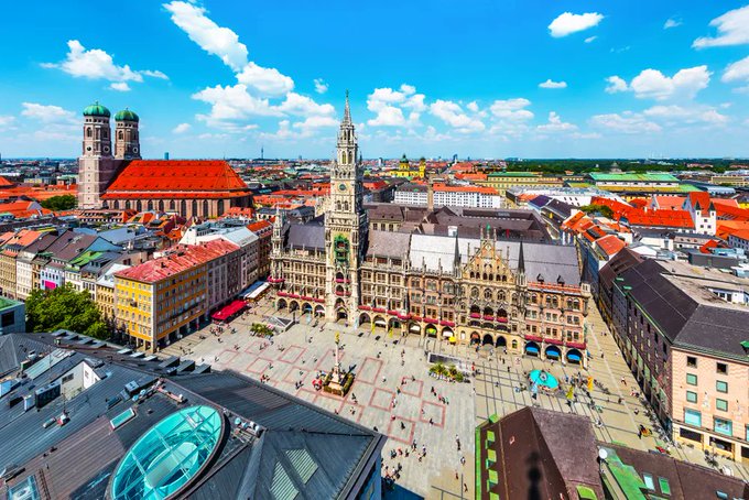 An aerial view of Marienplatz in Munich, Germany, showing the New Town Hall, the Frauenkirche cathedral with its iconic twin domed towers, and the bustling pedestrian square below.
