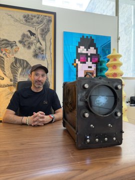 A man wearing a cap and black t-shirt sits at a wooden table next to a large, dark, studded cube-like object with a circular face/screen, with pixel art and a yellow pagoda visible in the background.