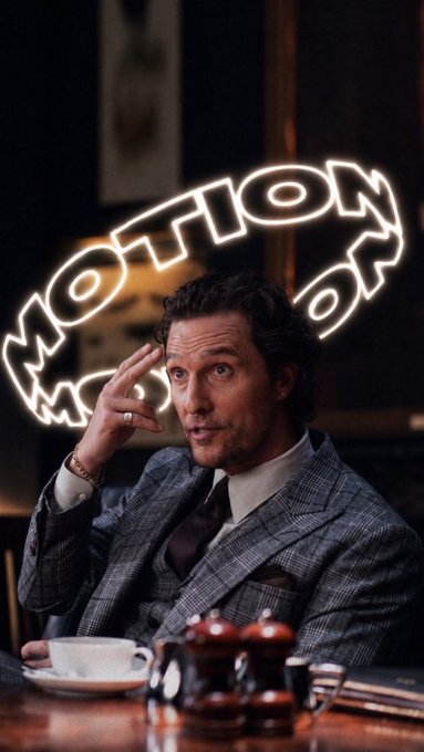 A man in a plaid suit sits at a table in front of a neon sign reading "MOTION," gesturing toward his head while speaking.