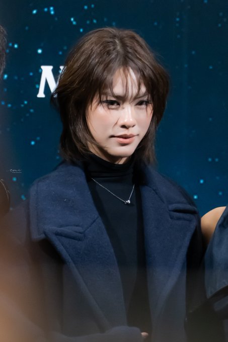 A young woman with a shoulder-length bob haircut wears a dark blue coat over a black turtleneck, posing in front of a blue backdrop.