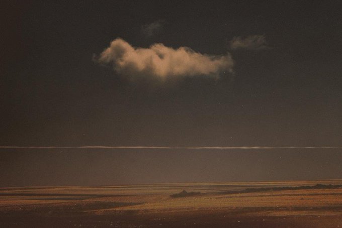 A dark, moody landscape shows a single, illuminated cloud floating above a flat horizon line that separates a deep sky from a shadowed foreground.