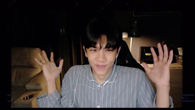 A young man in a striped shirt smiles at the camera while holding up both hands with palms facing forward, wearing white earphones in a dimly lit room with a sofa and lamp behind him.