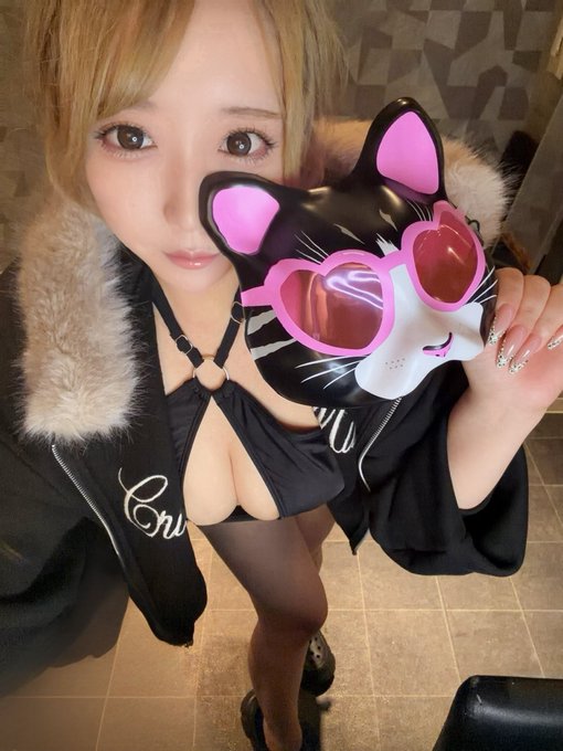 A selfie-style photo of a person with blonde hair wearing a black outfit with fur trim and a cut-out neckline. They hold a black cat-shaped mask with pink sunglasses close to the camera.