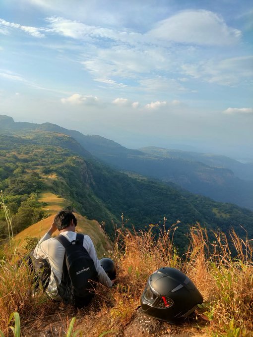 A person wearing a backpack and white shirt sits on a grassy cliff edge next to a motorcycle helmet, overlooking a vast green mountain valley under a partly cloudy sky.