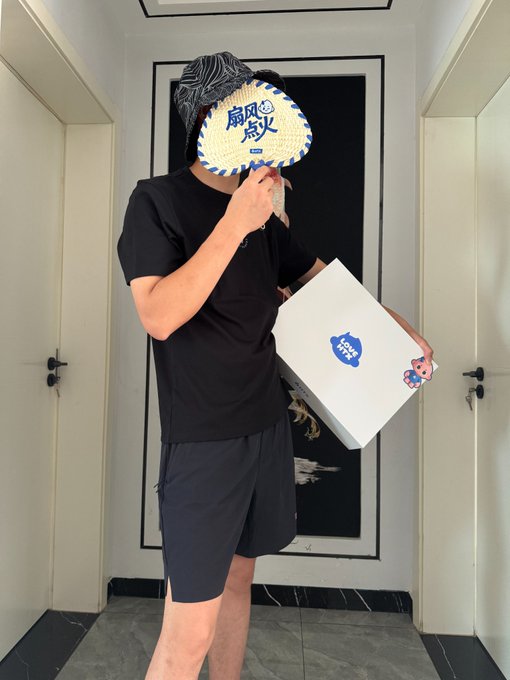 The image features a man holding up a white box with blue text, which appears to be a gift or present for someone else. The man is wearing black shorts and has a fan mask over his face. There are also two doors visible in the background, one on each side of the frame.