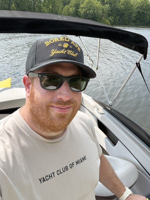 A red-bearded man wearing a 'Bored Ape Yacht Club' cap and 'Yacht Club of Miami' t-shirt takes a selfie while sitting on a boat.