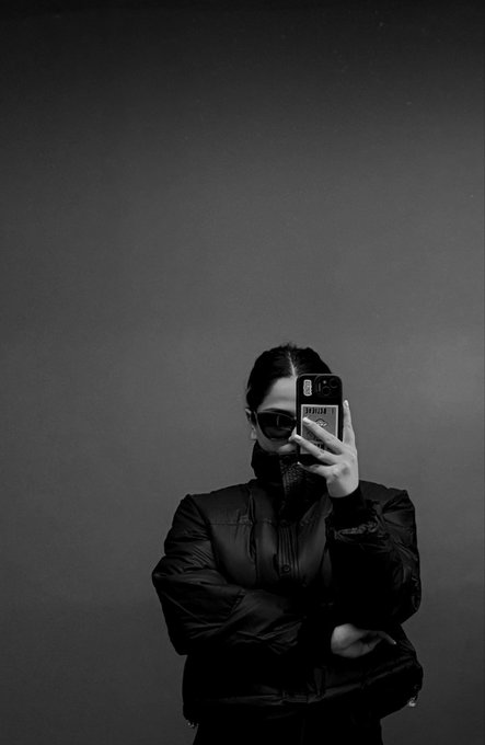 A person wearing sunglasses and a puffer jacket takes a black-and-white mirror selfie with their phone, obscuring their face. The minimalist composition, with vast empty space above them, reflects the introspective tone of the "Hello it's Moonaluna" introduction.