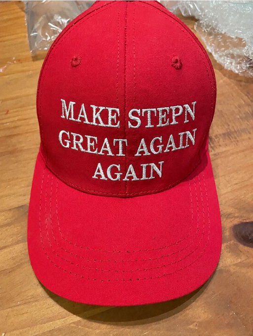 The image features a red baseball cap with white text that reads "MAKE STEPN GREAT AGAIN AGAIN". This message seems to be related to the ongoing political climate, as indicated by the caption provided. The cap appears to be sitting on top of a wooden table or countertop, suggesting it might have been placed there for display or storage purposes.