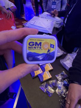 A hand holding a GM Mints tin labeled 'PROOF OF CONNECTIVITY', with more mints scattered on a table at what looks like a Web3 conference.