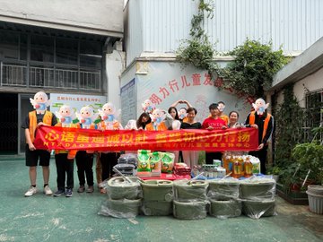 The image features a group of people standing together, holding up a large banner with Chinese characters that reads "旅行的最后一站是带我父母去参加 我老妈主动去买了箱旺仔牛奶". The group is composed of people in various positions, with some standing closer to the camera and others further away.

The scene takes place outdoors, possibly at a park or an event venue. There are several potted plants scattered throughout the area, adding greenery to the setting. A handbag can also be seen near one of the individuals in the group. The overall atmosphere appears to be festive and celebratory, with people gathered together for a special occasion or gathering.