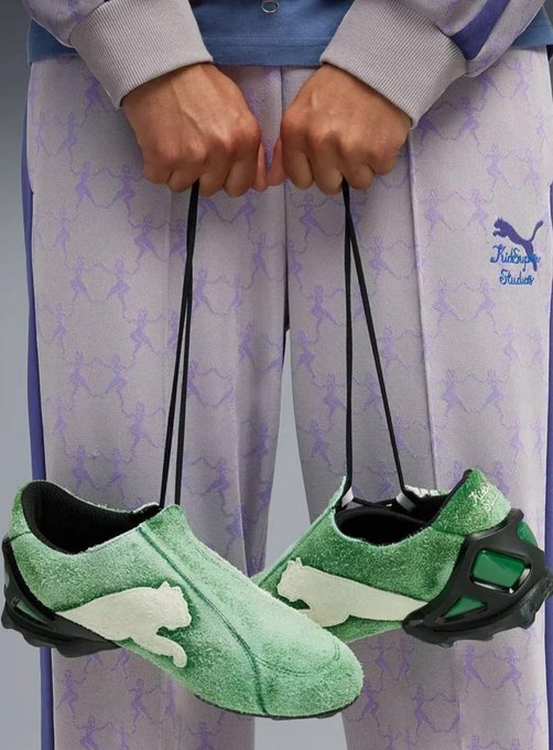 A person wearing purple patterned sweatpants holding a pair of green fuzzy Puma 'Waltniz' slippers by their laces.