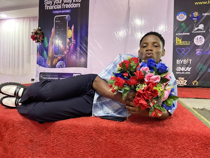 A young man reclining on a red carpet holding a large bouquet of red, pink, and blue roses, posing in front of sponsor banners including "Bybit" and a poster reading "Slay your way into financial freedom."