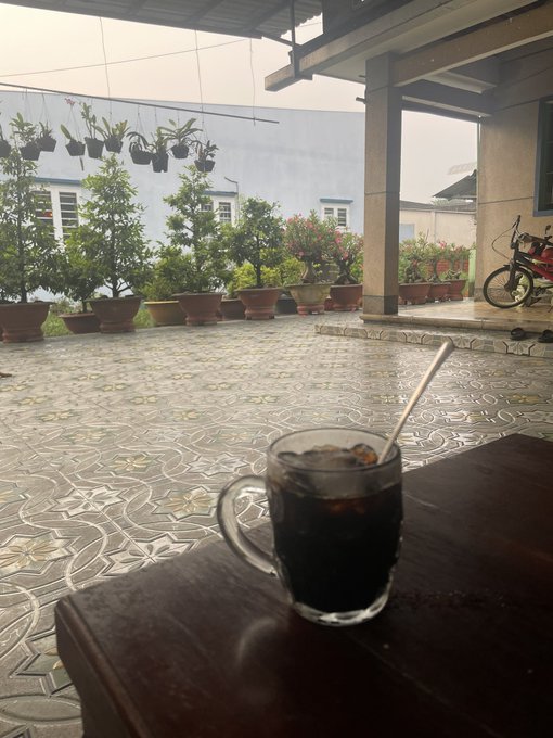 A glass of iced coffee with a straw sitting on a dark table, overlooking a tiled patio with potted plants, hanging orchids, and a bicycle in the background.