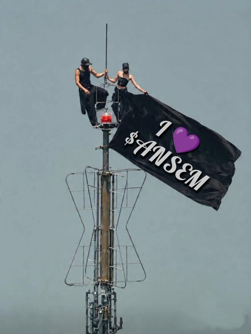 Two people stand atop a communications tower/spire, unfurling a black flag that reads "I [purple heart] $ANSCM" against a gray sky.