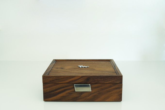 A rectangular wooden box with a visible grain pattern, a small metal handle on the front, and a decorative metal emblem on the lid, photographed against a white background.