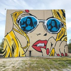 A large mural on a concrete wall depicts a stylized woman's face wearing white sunglasses, with two Mona Lisa portraits reflected in the lenses, complemented by yellow hair and red lips.