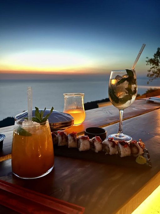 A sunset view overlooks cocktails and sushi on a waterfront table for a relaxed “Gm Degen.” spread. No other readable text, numbers, tickers, or names are visible.