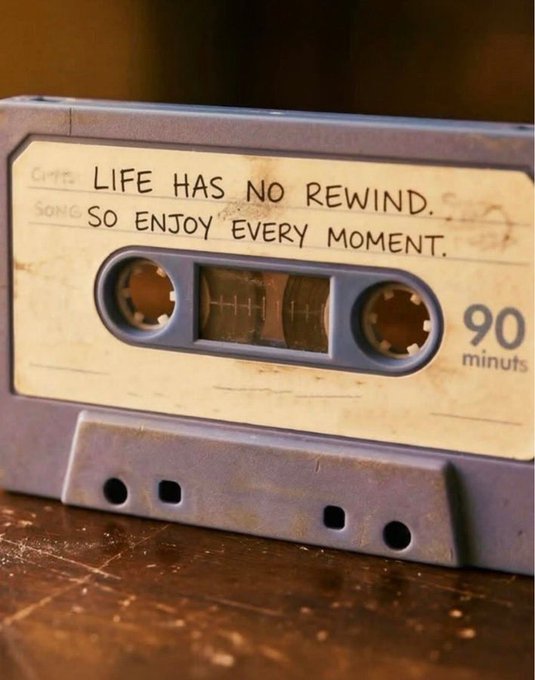 The image features an old, worn-out blue cassette tape with a quote written on it. The tape has two holes for the speaker, indicating its age or potential decline. The quote reads "Life Has No Wind. So Enjoy Every Moment." This message seems to be related to the concept of appreciating life's fleeting moments and finding joy in everyday experiences.
