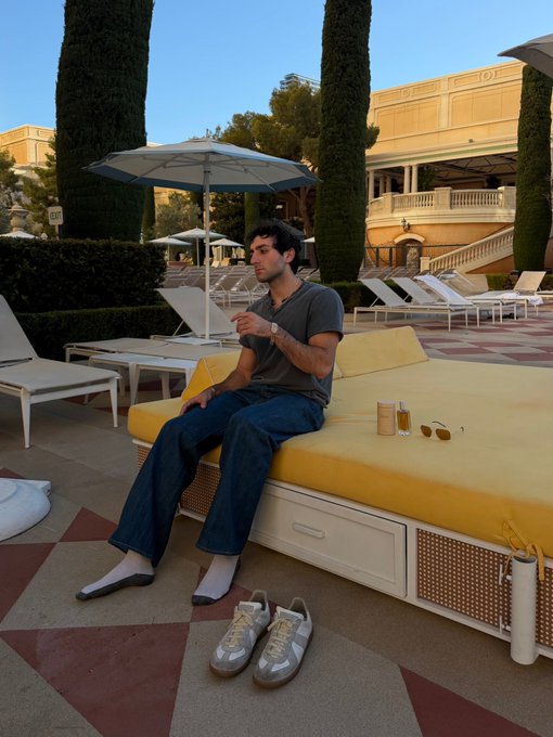 A man in a grey shirt and jeans lounges on a yellow daybed at an upscale resort, wearing designer sneakers placed beside him, evoking the post's joke about a casually dressed insider enjoying luxury after profiting from leaked information about the "cashcat" listing.