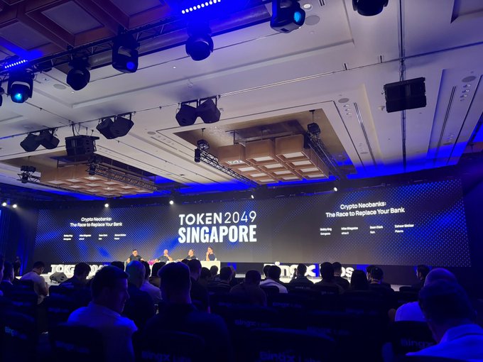 The image shows a panel discussion at the TOKEN2049 Singapore crypto conference, with the main screen displaying "Crypto Neobanks: The Race to Replace Your Bank." The accompanying post promotes a "BIGOD RWA AIRDROP" tied to real-world asset tokenization, connecting to the conference's focus on blockchain and crypto innovation shown in the scene.