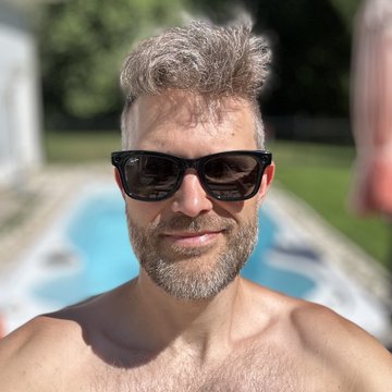 A close-up selfie of a shirtless man with graying hair and beard wearing sunglasses, with a pool visible in the background.