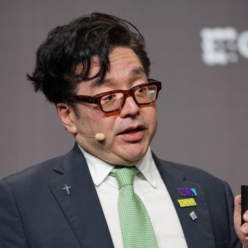 Tom Lee, a well-known market strategist, speaks at a podium while wearing a dark suit, green tie, and glasses, with a lapel pin visible on his jacket. The image relates to the accompanying post featuring his bullish prediction that Ethereum could surge 10x if it breaks the $5,000 price level after years of consolidation.