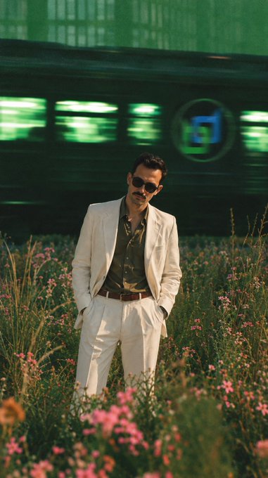 A man in a white suit and sunglasses standing in a field of pink flowers with a green-lit train passing in the background, creating a cinematic portrait.