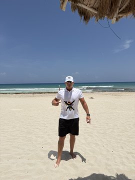 A man in a white t-shirt, black shorts, sunglasses and white cap stands on a sandy beach giving a thumbs-up, with the ocean and a thatched palapa roof behind him.