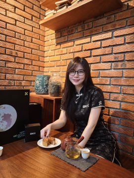 A young woman with glasses wearing a dark floral cheongsam dress sits at a wooden table in a brick-walled café, pouring tea from a glass pot alongside a plate of pastries.