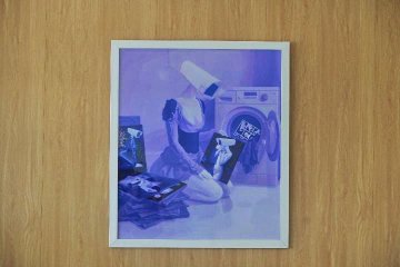 A framed print with a purple tint showing a woman with her head in a bag surrounded by cassette tapes, hung on a wooden wall.
