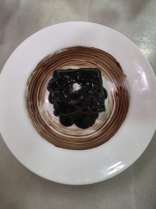 A top-down view of a white plate containing a square chocolate brownie topped with a dollop of chocolate sauce and powdered sugar, with chocolate sauce smeared in a circular pattern around it.