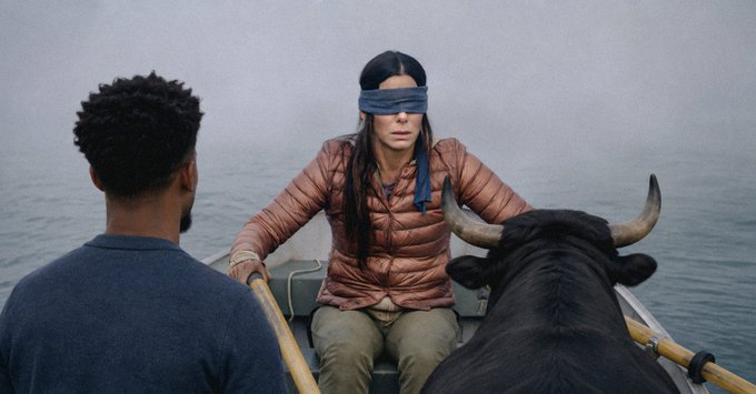 A scene from the show 'The OA' showing a woman with long dark hair wearing a blindfold, a rust-colored puffer jacket, and tan pants sitting in a boat with a black ox, while a man rows from behind.