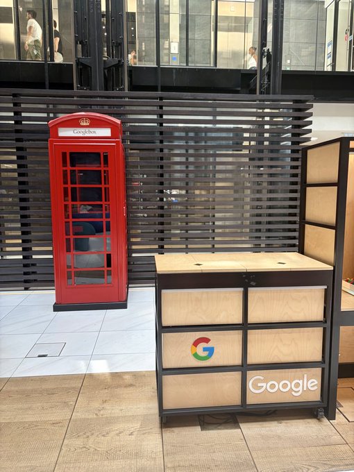 A red "Gosforth" British telephone booth stands next to a wooden Google-branded storage cabinet in what appears to be a Google office lobby. The image contrasts a traditional symbol of communication (the iconic phone booth) with a tech giant, visually emphasizing the bold claim that Zebec ($ZBCN) will surpass Google by positioning itself as the "Payments and Payroll of the future."