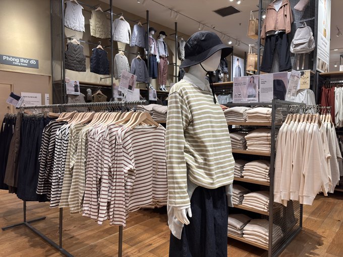 A retail clothing store display features a mannequin wearing a striped sweater and bucket hat, surrounded by racks of neutral-toned garments and a 'Phòng thử đồ' (Fitting Rooms) sign.