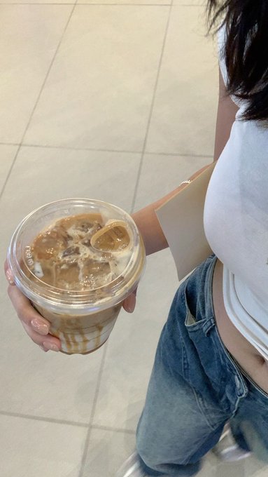 A woman in a white tank top and jeans holds an iced coffee while standing on a tiled floor. The relaxed, casual scene captures a moment of taking a coffee break before diving into work, fitting the post's message about slowing down, enjoying a Sunday morning, and prioritizing a clear mind over constant market activity.