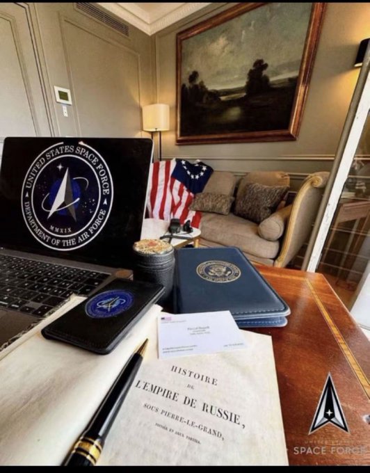 A workspace scene featuring a laptop with the United States Space Force logo, an American flag, a book titled 'HISTOIRE DE L'EMPIRE DE RUSSIE' under Pierre-le-Grand, and various official items.