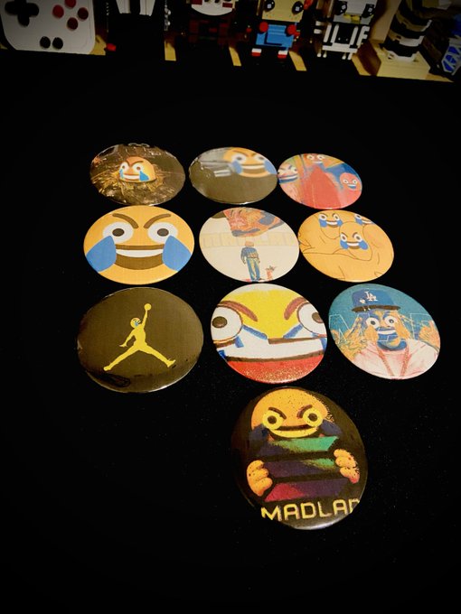 A photograph showing ten circular pin badges or buttons arranged on a black surface, featuring various cartoon faces, sports logos (including a yellow 'Jumpman' logo), and characters, with one button at the bottom reading 'MADLAB'.