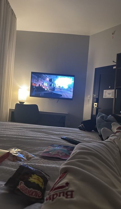 A view from a hotel bed showing a person in Budweiser sweatpants lounging with snacks including a bag of Swad chips and a Pokémon card, watching TV in a room with gray walls.