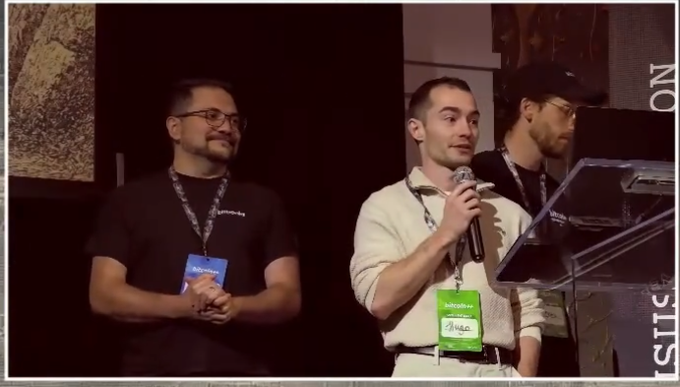 Three men on stage at a tech event, with the central figure speaking into a microphone while wearing conference badges labeled 'SPEAKER.'