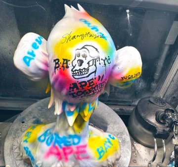 The image features a cake that has been decorated with graffiti art, including the words "Bored Ape" written on its head. The cake appears to be made for an event or celebration related to the Asia Tour, as indicated by the presence of the word "Asia" in one of the descriptions. The cake is placed on a table and there are two other cakes visible in the background.