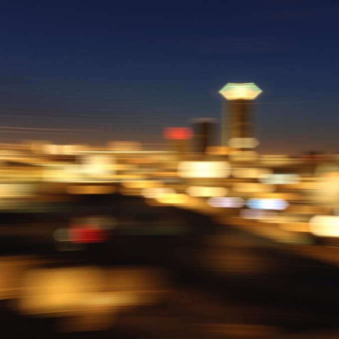 A heavily motion-blurred nighttime cityscape showing streaks of car lights and an illuminated tower against a dark blue sky.