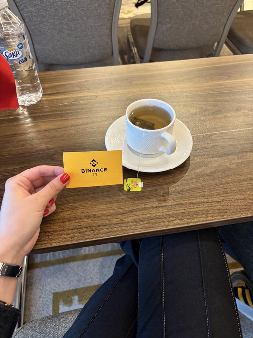The image shows a person's hand holding up a yellow business card with the word "Binance" written on it, along with a cup of tea sitting on top of a wooden table. The background includes chairs and other tables, suggesting that this scene takes place in an office or workspace setting.