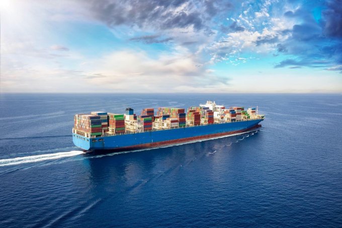 An aerial view of a large blue container ship loaded with multicolored shipping containers, sailing across a calm blue ocean under a partly cloudy sky.