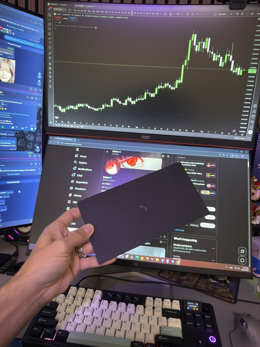 A person holds up a sleek black envelope addressed to "MissionGains" in front of their trading setup featuring a candlestick chart on the top monitor and the X platform on the bottom screen, with their custom keyboard visible below—capturing the exciting moment of unboxing their new X Money debit card while in their trading workspace.
